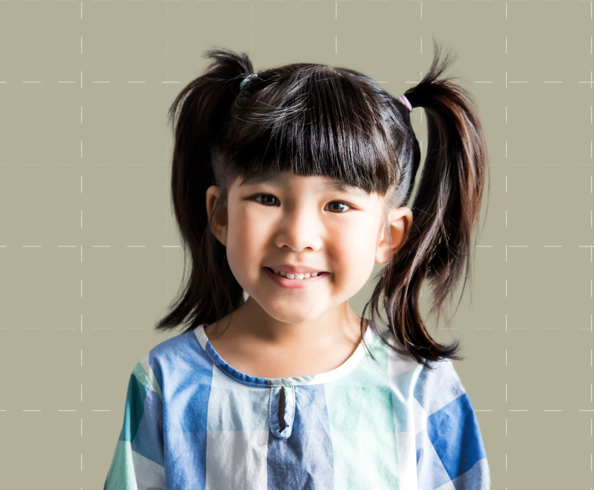Young girl with dark hair in pigtails, wearing a blue and white checkered shirt, smiling in front of a plain light background.