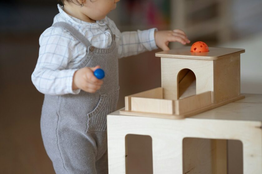A young child in grey overalls plays with a wooden toy, holding a blue ball and reaching for an orange ball on top of the toy structure.