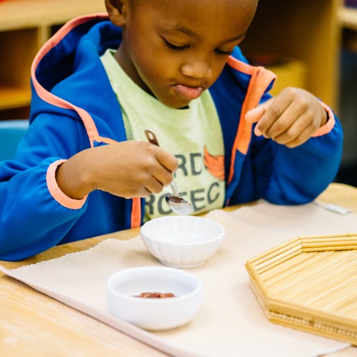 A child sits at a table using a spoon to scoop from a white bowl, with another bowl and a wooden tray on the table.