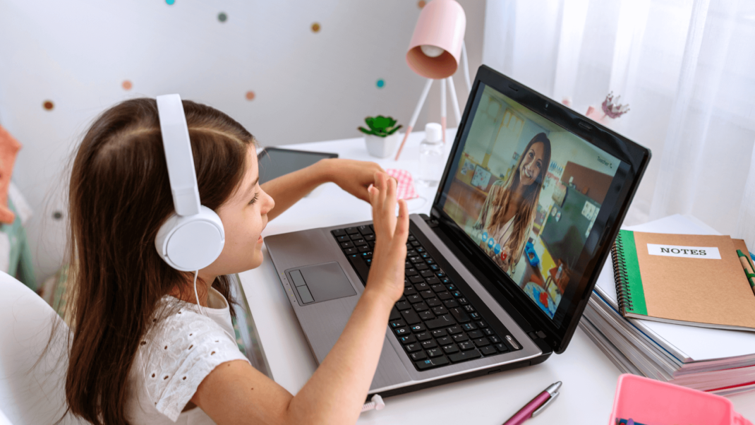 A young girl wearing headphones sits at a desk, using a laptop to video chat with a woman. Notebooks, a lamp, and other desk items are visible.