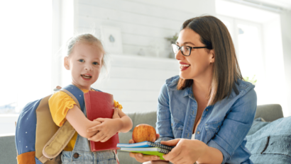 A young girl with a backpack holds a red book while an adult woman hands her notebooks and an apple in a bright room.