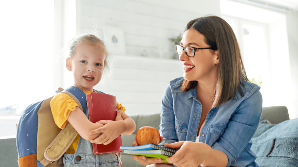 A young girl with a backpack holds a red book while an adult woman hands her notebooks and an apple in a bright room.