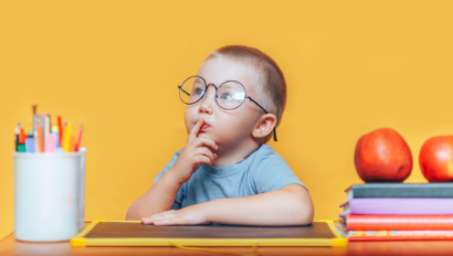 A young child wearing glasses sits at a desk with a finger on their lips, surrounded by books, apples, and colored pencils, against a plain orange background.