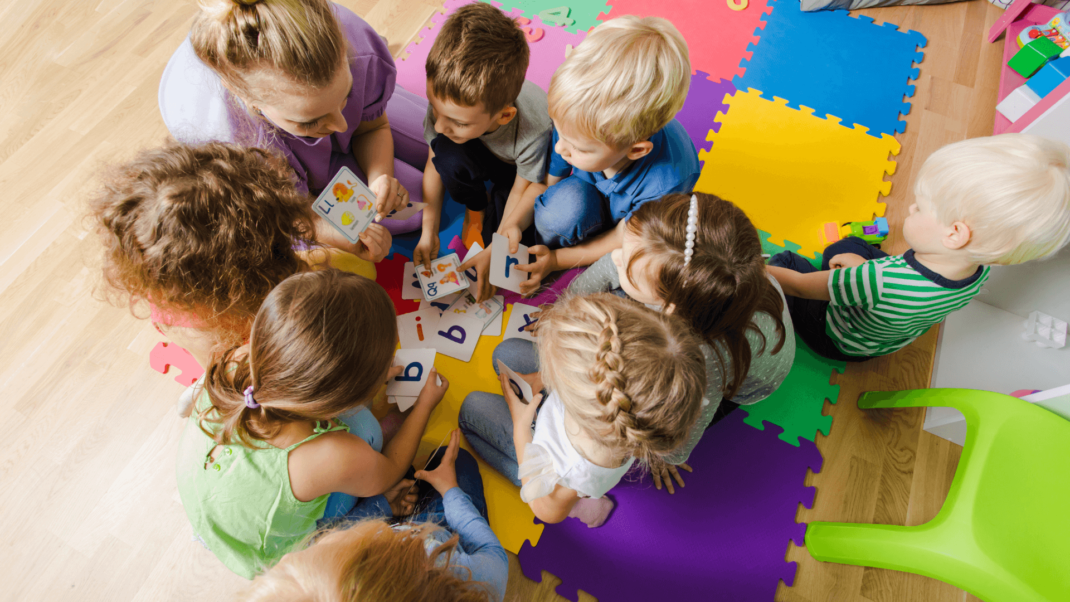 A group of young children sits in a circle on colorful foam mats, looking at and holding alphabet cards, guided by an adult.