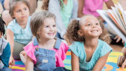 A group of young children sit on a colorful mat, smiling and looking toward a person reading a book to them.