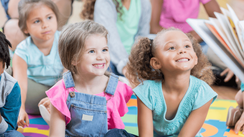 A group of young children sit on a colorful mat, smiling and looking toward a person reading a book to them.