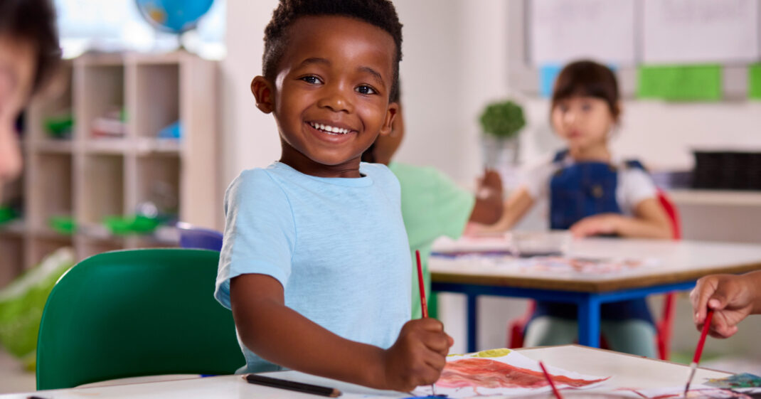 A young boy smiles while painting at a table in a classroom, with other children working in the background.