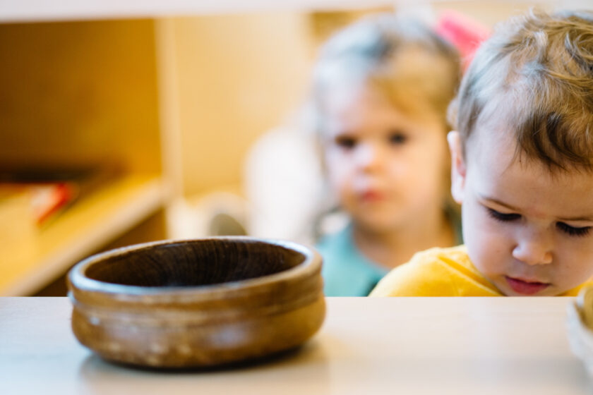 Two young children stand behind a wooden bowl on a table; one child is in focus and facing downward, while the other appears blurred in the background.