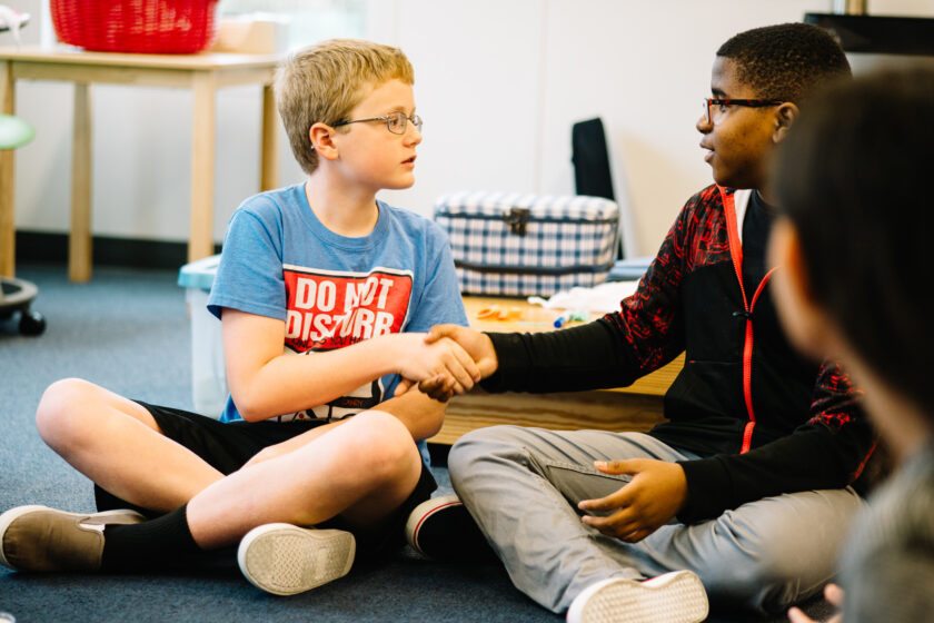 Two boys sit cross-legged on the floor and shake hands in a classroom setting, with another student partially visible in the foreground.